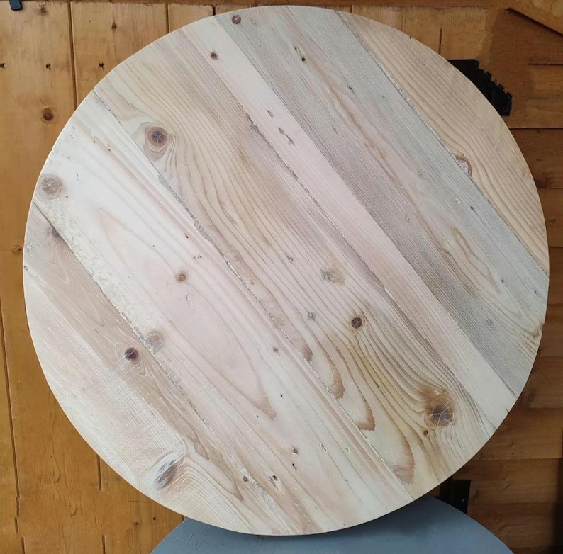 Op de afbeelding: Een ronde houten tafelblad gemaakt van gerecycled hout. Het hout heeft een natuurlijke lichtbruine kleur met wat donkerdere bruine knopen en nerven.