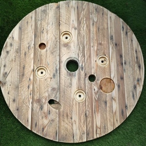 May include: A large, round wooden spool with a weathered, rustic finish. The spool has several circular holes and a large, rectangular hole in the centre. The spool is made of natural wood with a light brown colour.