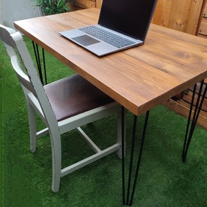 May include: A wooden desk with a light brown finish and black hairpin legs. A laptop computer is on the desk. The desk is in front of a green artificial grass background.