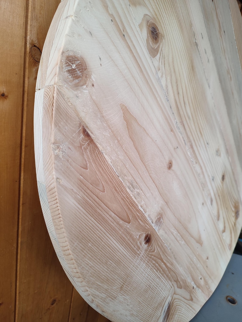 Op de afbeelding: Een close-up van een licht houten ovaal tafelblad. De houtnerf is zichtbaar en het oppervlak is glad.