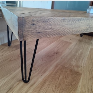 May include: A reclaimed wood coffee table featuring a natural, weathered tabletop and black hairpin legs. The table's surface displays visible wood grain and nail heads. The black hairpin legs offer a modern contrast to the table's rustic aesthetic. The table is approximately 120 cm long.