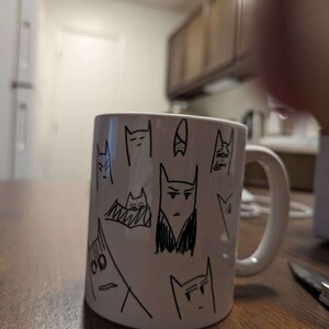May include: White ceramic mug with black line drawings of cats on it. The cats have different expressions and are drawn in a simple style.