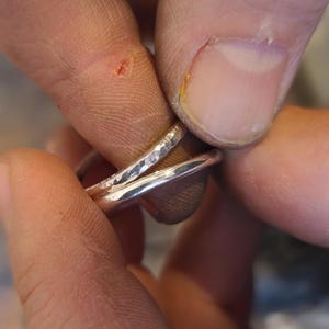 Può includere: Due anelli d'argento, uno con una texture martellata e l'altro liscio, sono tenuti insieme dalle dita.