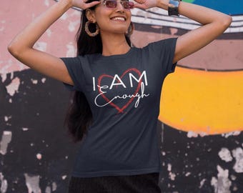 May include: A navy blue t-shirt with the words "I AM enough" in white script and a red heart design. The shirt is short-sleeved and has a crew neck. The background is a colorful mural.