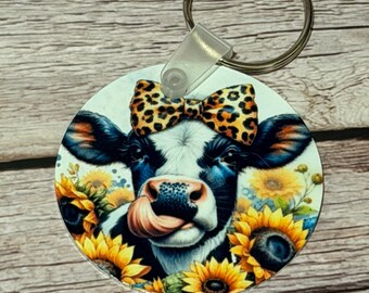 May include: A round keychain featuring a cartoon cow with a black and white face, wearing a leopard print bow, and surrounded by sunflowers. The cow is sticking out its tongue. The keychain has a silver ring.