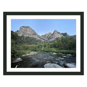 May include: A scenic view of a river flowing through a valley with two rocky mountains in the background. The river is surrounded by lush green trees and vegetation.
