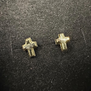 May include: A pair of small, gold-colored cross stud earrings. The crosses have a textured design and are set against a dark background. These earrings are a religious-themed accessory.