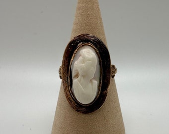 May include: A gold ring with a cameo featuring a carved white profile of a woman. The cameo is set in an oval frame with intricate details.