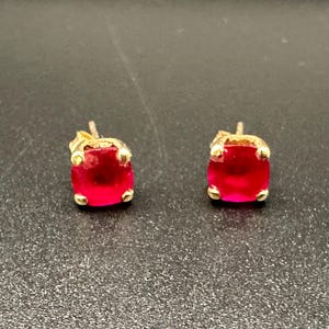 May include: A pair of stud earrings featuring round, vibrant red gemstones set in gold-toned metal prongs. Each earring has a classic four-prong setting that secures the circular red stone, creating a simple and elegant design for these jewellery pieces.