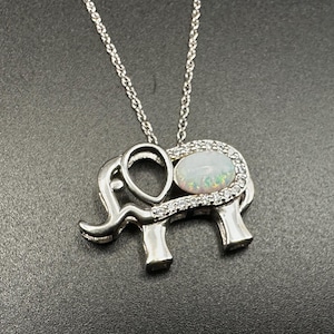 May include: Silver elephant pendant necklace with an oval opal gemstone center, surrounded by small clear stones. The elephant design has detailed features and is suspended from a delicate chain.