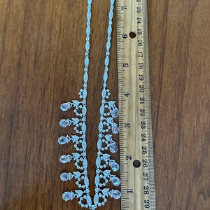 May include: A silver-toned necklace with clear and pink teardrop-shaped pendants. The necklace is approximately 16 inches long and is displayed on a wooden surface next to a ruler.