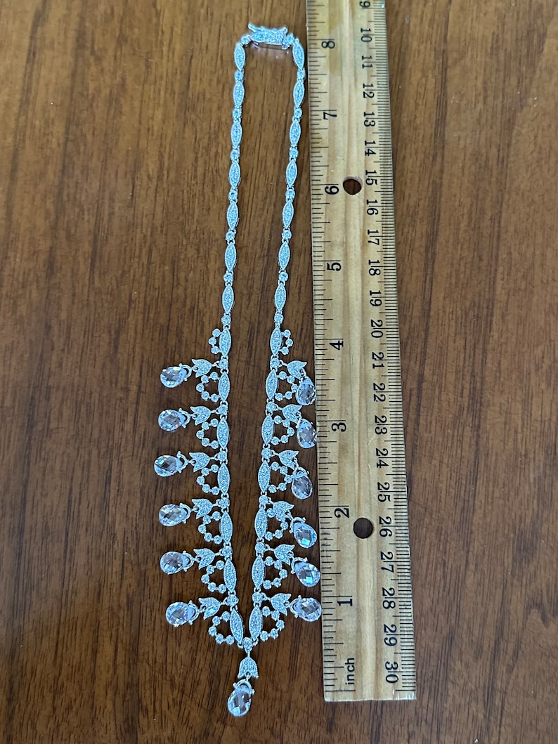 May include: A silver-toned necklace with clear and pink teardrop-shaped pendants. The necklace is approximately 16 inches long and is displayed on a wooden surface next to a ruler.