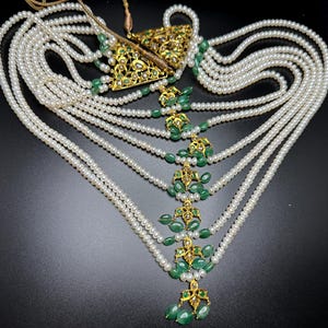 Puede incluir: Un collar elaborado con m&uacute;ltiples hilos de perlas blancas. El collar est&aacute; adornado con elementos de filigrana dorados y gemas verdes vibrantes. El dise&ntilde;o incluye un colgante triangular y gotas decorativas.