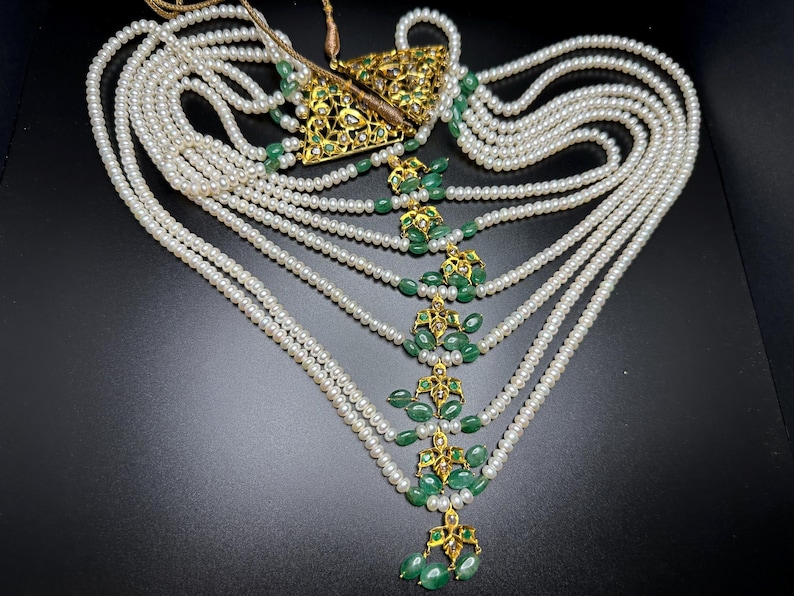 Puede incluir: Un collar elaborado con m&uacute;ltiples hilos de perlas blancas. El collar est&aacute; adornado con elementos de filigrana dorados y gemas verdes vibrantes. El dise&ntilde;o incluye un colgante triangular y gotas decorativas.
