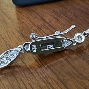 May include: Close-up of a silver bracelet with a decorative clasp. The bracelet features a series of marquise-shaped links, each set with small, clear gemstones. The clasp is rectangular with the text "925" and "FZS" engraved on it.
