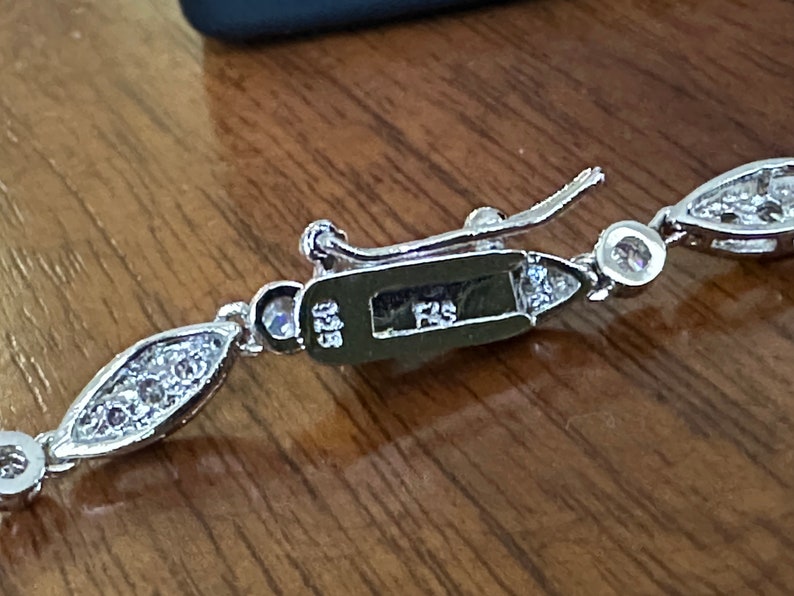 May include: Close-up of a silver bracelet with a decorative clasp. The bracelet features a series of marquise-shaped links, each set with small, clear gemstones. The clasp is rectangular with the text "925" and "FZS" engraved on it.