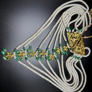 Puede incluir: Un tocado ornamentado con m&uacute;ltiples hilos de perlas blancas. El dise&ntilde;o incorpora detalles de metal dorado y piedras preciosas de color verde esmeralda. El tocado tiene forma triangular y est&aacute; sobre un fondo oscuro.