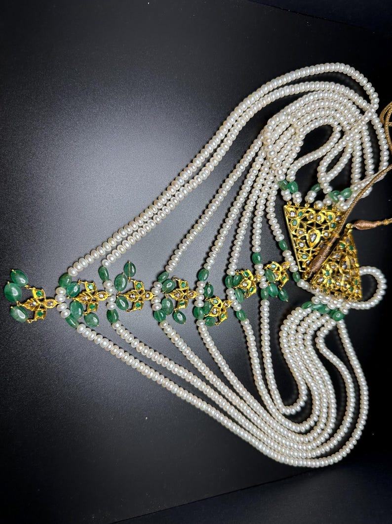 Puede incluir: Un tocado ornamentado con m&uacute;ltiples hilos de perlas blancas. El dise&ntilde;o incorpora detalles de metal dorado y piedras preciosas de color verde esmeralda. El tocado tiene forma triangular y est&aacute; sobre un fondo oscuro.