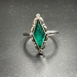 Puede incluir: Un anillo de plata con una piedra preciosa verde engastada en un engaste en forma de diamante. El anillo est&aacute; adornado con intrincados detalles alrededor de la piedra preciosa.