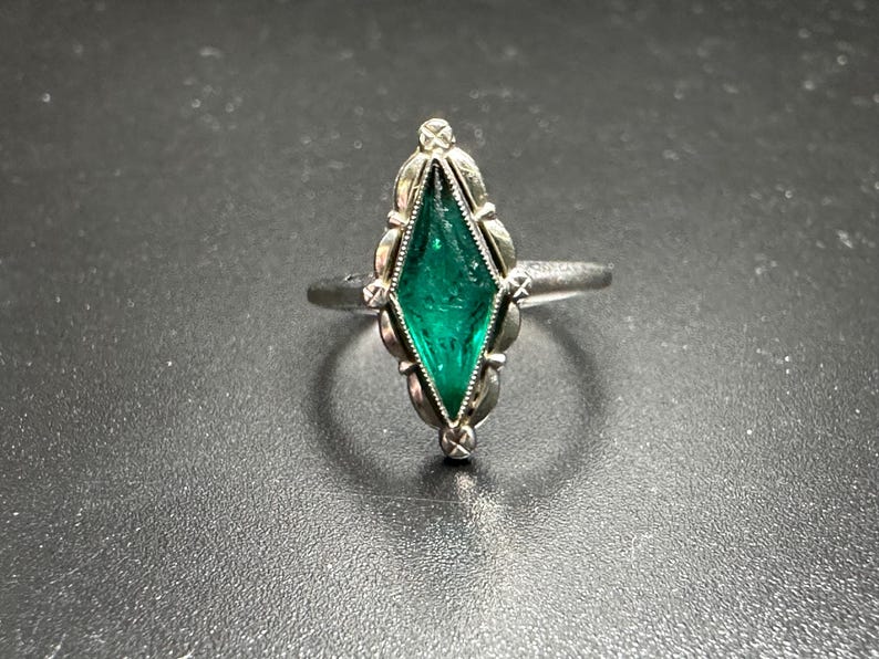 Puede incluir: Un anillo de plata con una piedra preciosa verde engastada en un engaste en forma de diamante. El anillo est&aacute; adornado con intrincados detalles alrededor de la piedra preciosa.