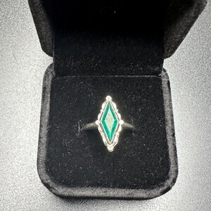 Puede incluir: Un anillo de plata con una piedra preciosa verde en un engaste en forma de diamante. El anillo est&aacute; en una caja de anillo de terciopelo negro.