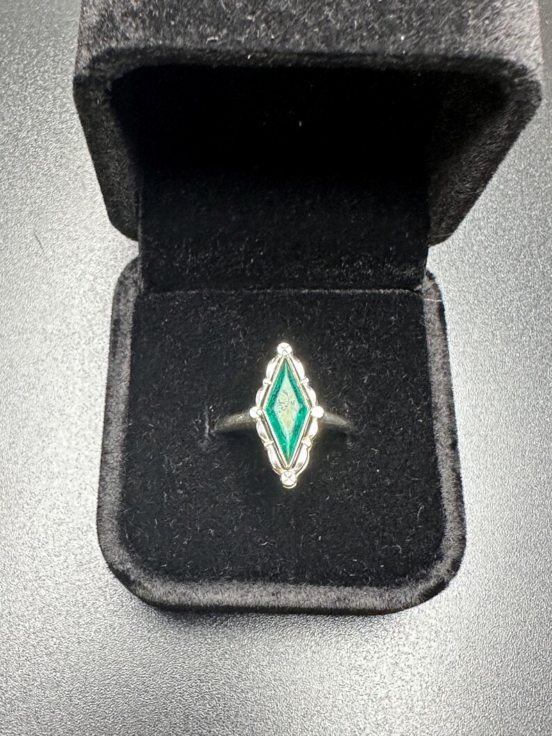 Puede incluir: Un anillo de plata con una piedra preciosa verde en un engaste en forma de diamante. El anillo est&aacute; en una caja de anillo de terciopelo negro.