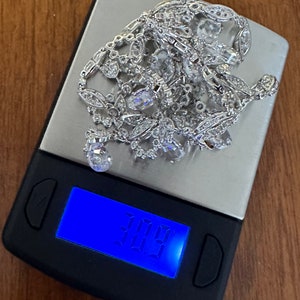 May include: A silver necklace with clear gemstones and intricate detailing rests on a digital scale. The scale displays a weight of 30.9, suggesting the necklace's weight in an unknown unit. The background is a wooden surface.