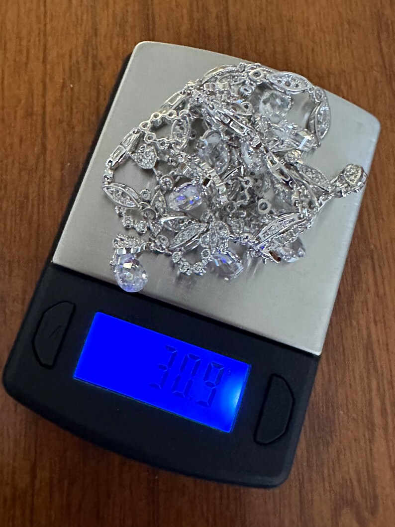 May include: A silver necklace with clear gemstones and intricate detailing rests on a digital scale. The scale displays a weight of 30.9, suggesting the necklace's weight in an unknown unit. The background is a wooden surface.
