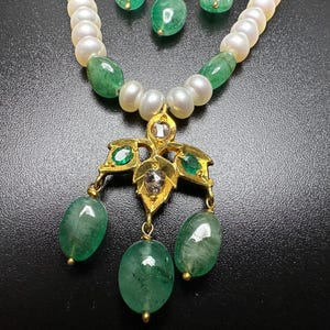 Puede incluir: Un conjunto de collar y pendientes con una cadena de perlas blancas y esmeraldas verdes. El colgante es dorado con dise&ntilde;os en forma de hoja y detalles de esmeraldas, con tres gotas de esmeralda colgantes. Los pendientes tienen gotas de esmeralda.