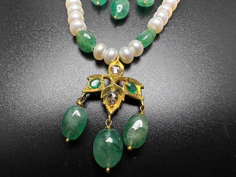 Puede incluir: Un conjunto de collar y pendientes con una cadena de perlas blancas y esmeraldas verdes. El colgante es dorado con dise&ntilde;os en forma de hoja y detalles de esmeraldas, con tres gotas de esmeralda colgantes. Los pendientes tienen gotas de esmeralda.