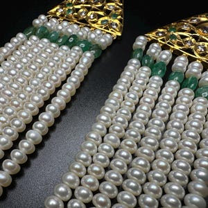 Puede incluir: Primer plano de dos pendientes ornamentados con perlas y piedras preciosas. Cada pendiente presenta m&uacute;ltiples hilos de perlas blancas, acentuados con piedras preciosas verdes y detalles de filigrana dorada.
