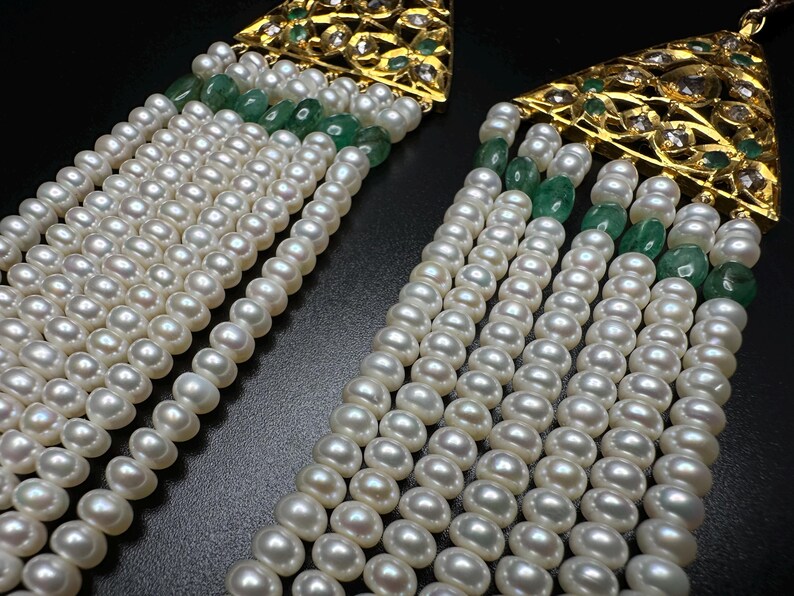 Puede incluir: Primer plano de dos pendientes ornamentados con perlas y piedras preciosas. Cada pendiente presenta m&uacute;ltiples hilos de perlas blancas, acentuados con piedras preciosas verdes y detalles de filigrana dorada.