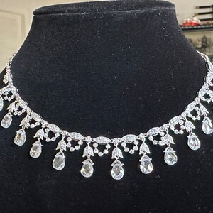 May include: A silver-toned necklace with a delicate design. The necklace features a series of clear, teardrop-shaped pendants suspended from a detailed chain. The chain is adorned with small, sparkling accents, creating an elegant and eye-catching piece.