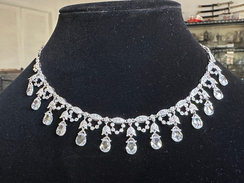 May include: A silver-toned necklace with a delicate design. The necklace features a series of clear, teardrop-shaped pendants suspended from a detailed chain. The chain is adorned with small, sparkling accents, creating an elegant and eye-catching piece.