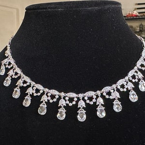 May include: A silver-toned necklace with clear, teardrop-shaped pendants. The necklace features a delicate design with small, sparkling accents and is displayed on a black surface. The jewelry is likely for special occasions.