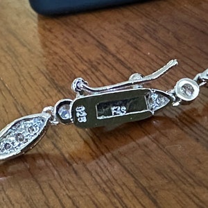 May include: Close-up of a silver bracelet with a decorative clasp. The bracelet features a series of bezel-set, round and marquise-shaped clear stones. The clasp is engraved with "925" and "FS".