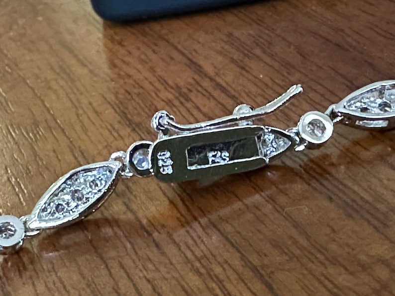 May include: Close-up of a silver bracelet with a decorative clasp. The bracelet features a series of bezel-set, round and marquise-shaped clear stones. The clasp is engraved with "925" and "FS".