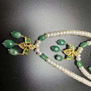 Puede incluir: Primer plano de un conjunto de collar y pendientes de dos hilos. El collar presenta perlas blancas y esmeraldas verdes. Los pendientes y colgantes del collar tienen un dise&ntilde;o floral dorado con detalles de esmeraldas y diamantes. El fondo es negro.