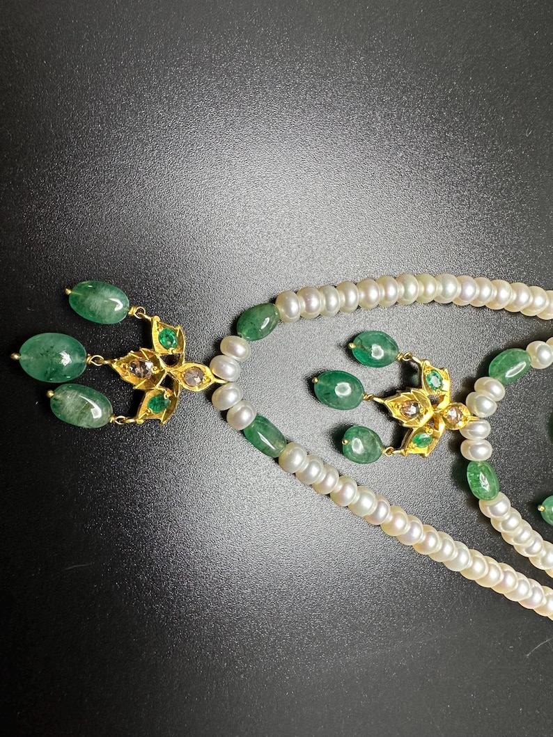 Puede incluir: Primer plano de un conjunto de collar y pendientes de dos hilos. El collar presenta perlas blancas y esmeraldas verdes. Los pendientes y colgantes del collar tienen un dise&ntilde;o floral dorado con detalles de esmeraldas y diamantes. El fondo es negro.