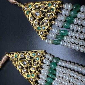 Puede incluir: Primer plano de dos collares de oro con intrincados colgantes triangulares. Los colgantes est&aacute;n adornados con diamantes y esmeraldas. Cada collar presenta m&uacute;ltiples hilos de perlas blancas y cuentas de esmeralda verde, creando un dise&ntilde;o lujoso y elegante.