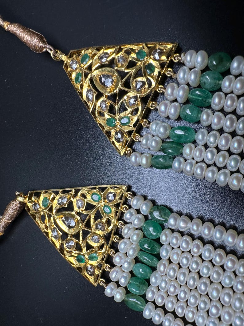 Puede incluir: Primer plano de dos collares de oro con intrincados colgantes triangulares. Los colgantes est&aacute;n adornados con diamantes y esmeraldas. Cada collar presenta m&uacute;ltiples hilos de perlas blancas y cuentas de esmeralda verde, creando un dise&ntilde;o lujoso y elegante.