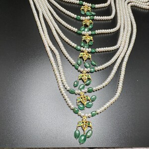 Puede incluir: Un collar de perlas de m&uacute;ltiples hilos con detalles de esmeralda y oro. El collar presenta m&uacute;ltiples hilos de peque&ntilde;as perlas blancas. Colgantes florales dorados con piedras verdes esmeralda en forma de l&aacute;grima est&aacute;n espaciados uniformemente.