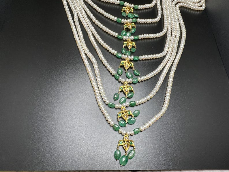 Puede incluir: Un collar de perlas de m&uacute;ltiples hilos con detalles de esmeralda y oro. El collar presenta m&uacute;ltiples hilos de peque&ntilde;as perlas blancas. Colgantes florales dorados con piedras verdes esmeralda en forma de l&aacute;grima est&aacute;n espaciados uniformemente.