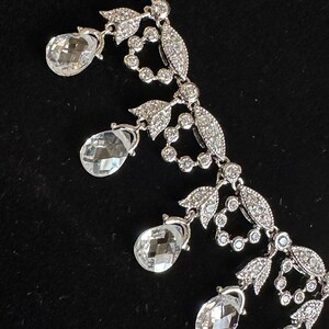 May include: A silver-toned necklace with clear, teardrop-shaped pendants and intricate floral and leaf detailing. The necklace features small, round, and sparkling accents throughout the design, creating a delicate and elegant appearance.