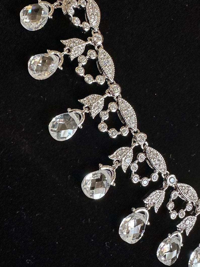 May include: A silver-toned necklace with clear, teardrop-shaped pendants and intricate floral and leaf detailing. The necklace features small, round, and sparkling accents throughout the design, creating a delicate and elegant appearance.