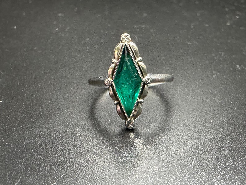 Puede incluir: Un anillo de plata con una piedra preciosa verde engastada en un engaste en forma de diamante. El anillo tiene un dise&ntilde;o delicado y ornamentado.