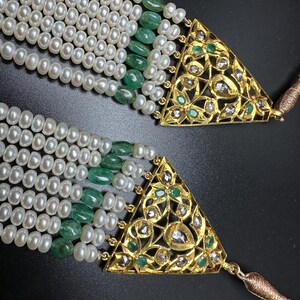 Puede incluir: Primer plano de un collar de perlas de varias hebras con cuentas de esmeralda y cierres triangulares dorados. Los cierres est&aacute;n intrincadamente dise&ntilde;ados con patrones florales y peque&ntilde;as piedras preciosas. El collar tiene una apariencia lujosa y elegante.