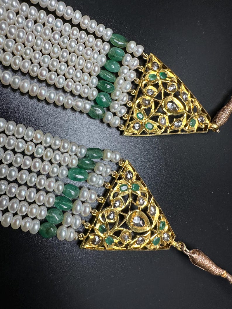 Puede incluir: Primer plano de un collar de perlas de varias hebras con cuentas de esmeralda y cierres triangulares dorados. Los cierres est&aacute;n intrincadamente dise&ntilde;ados con patrones florales y peque&ntilde;as piedras preciosas. El collar tiene una apariencia lujosa y elegante.
