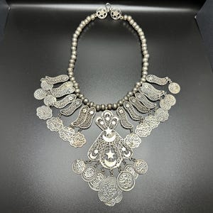 May include: A silver filigree necklace with a crescent moon and star pendant. The necklace is adorned with multiple small, round charms and has a beaded chain.