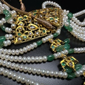 Puede incluir: Un collar ornamentado con m&uacute;ltiples hilos de perlas blancas, intercaladas con piedras preciosas verdes. Un colgante dorado con detalles intrincados y gemas incrustadas es el punto focal. El collar se presenta sobre un fondo oscuro.